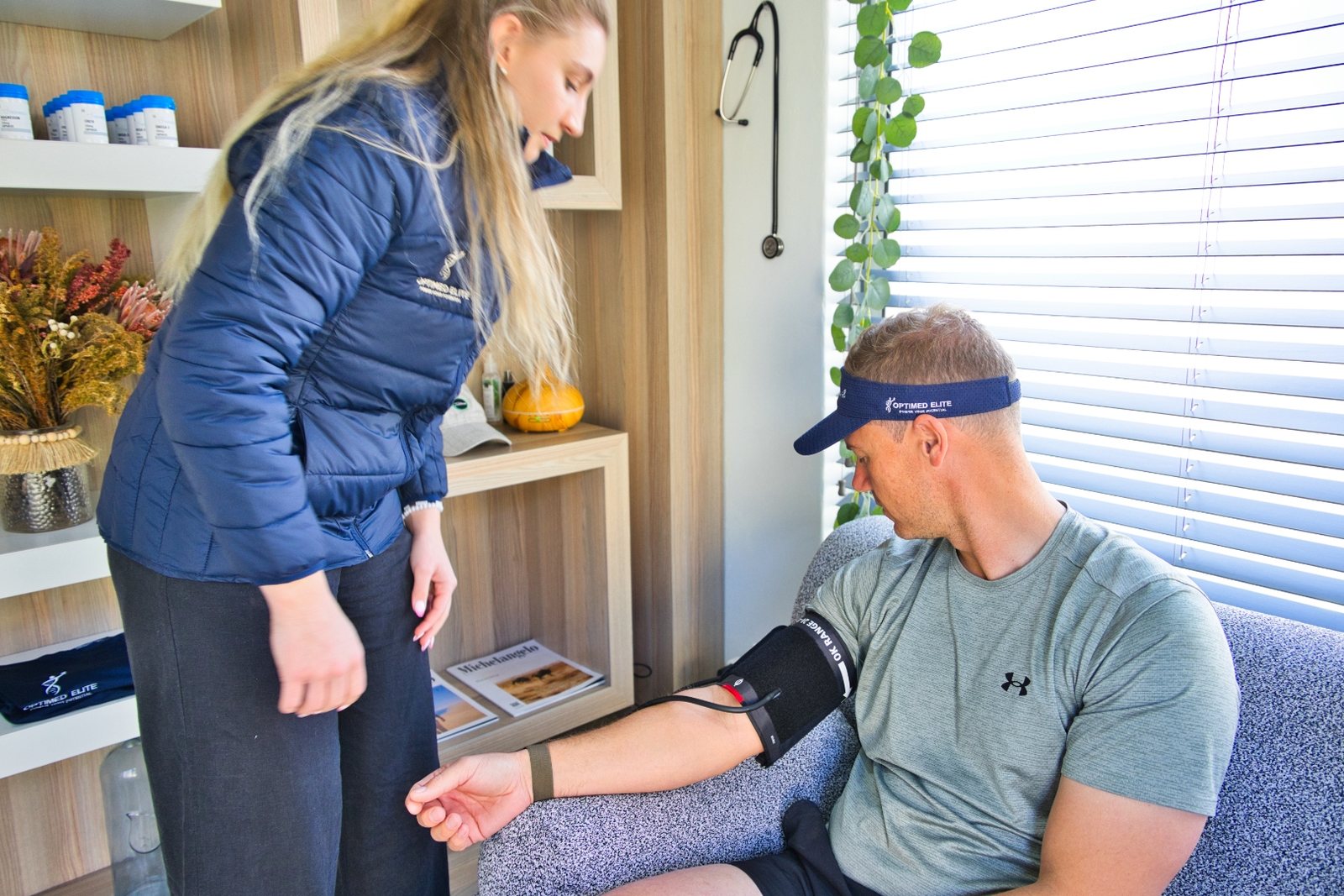 A clinician in an OptiMED Elite jacket fitting an upper-arm blood-pressure cuff to a seated patient in the consulting room; the patient wears an OptiMED Elite visor.