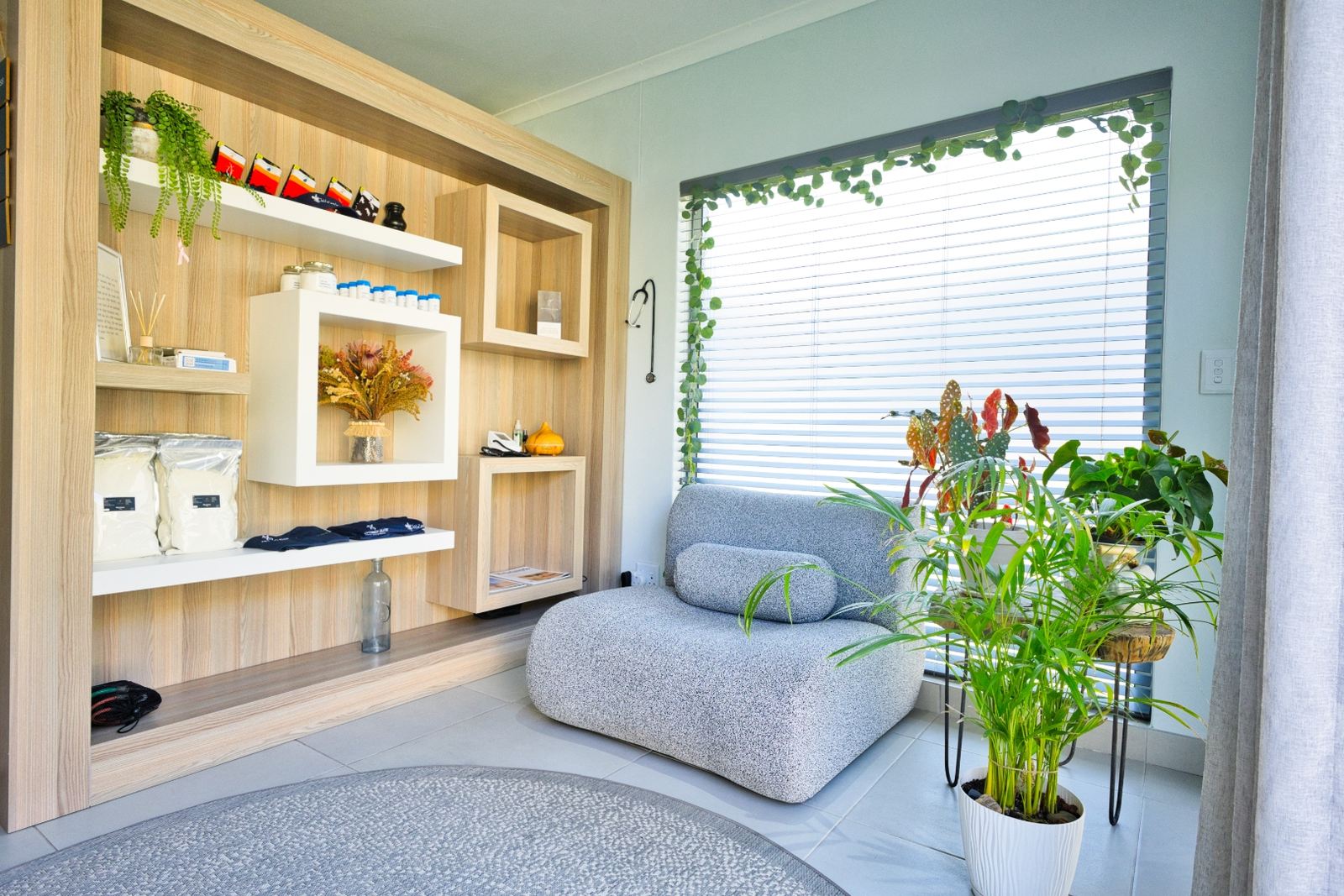 A quiet corner of the practice in Windhoek — a pale grey bouclé armchair beside a shuttered window, with built-in shelving holding supplement pouches, folded branded shirts, a reed diffuser and a dried protea arrangement, and potted plants alongside.