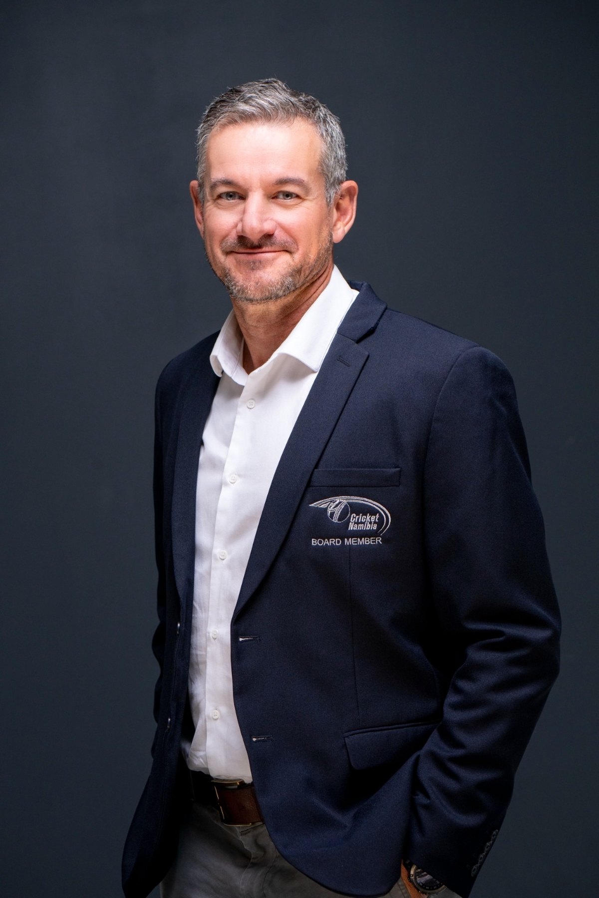 Dr Rudie van Vuuren, standing against a dark grey backdrop in a navy blazer and open-collar white shirt, hands in pockets.