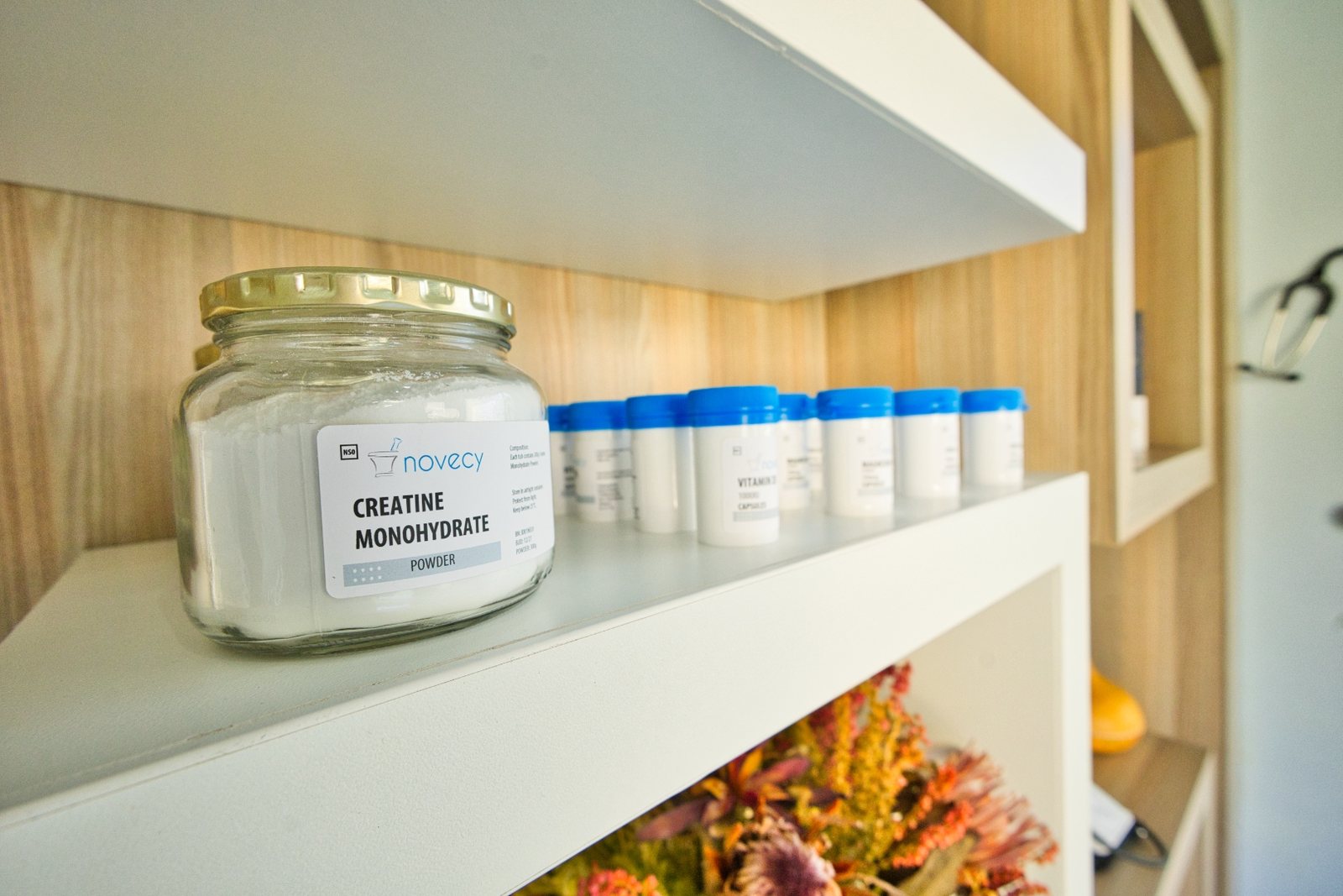 A glass jar of creatine monohydrate powder and a row of white, blue-capped supplement and vitamin bottles on the practice's white shelving.